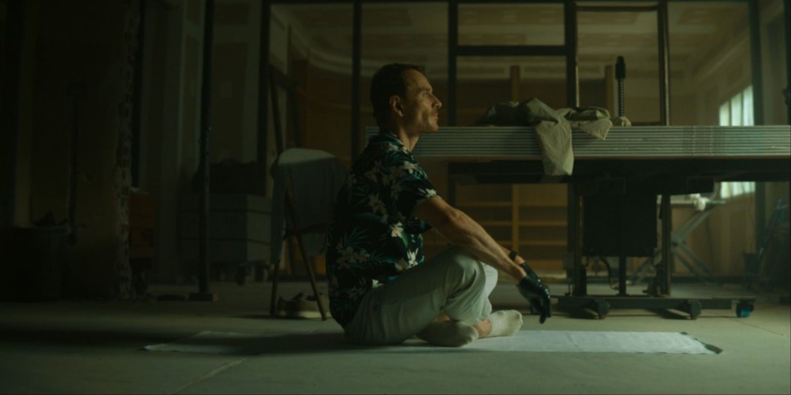 Michael Fassbender sitting on the floor criss-cross with black gloves on in The Killer.