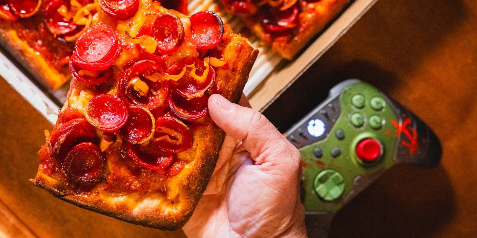 A person holding a slice of pizza with an Xbox controller in the background